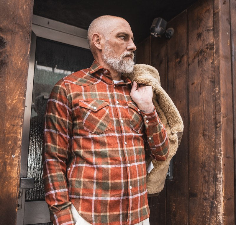 Mann in kariertem Western-Flanellhemd, Jacke mit Lammfellkragen über der Schulter, vor einem Holztor. Foto @chesapeakesofficial