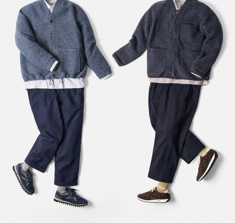 Zwei flach ausgelegte Outfits von Universal Works: Wollcardigans in Dunkel- und Hellblau, weißes Hemd, blaue Hose und Sneaker.